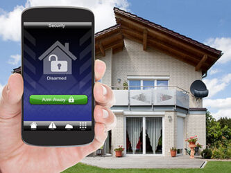 Hand holding a smartphone displaying a home security app with arm and disarm options, in front of a modern two-storey house.