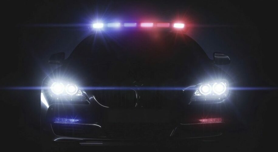 Front view of a police car at night with bright headlights on and flashing red and blue emergency lights across the roof.