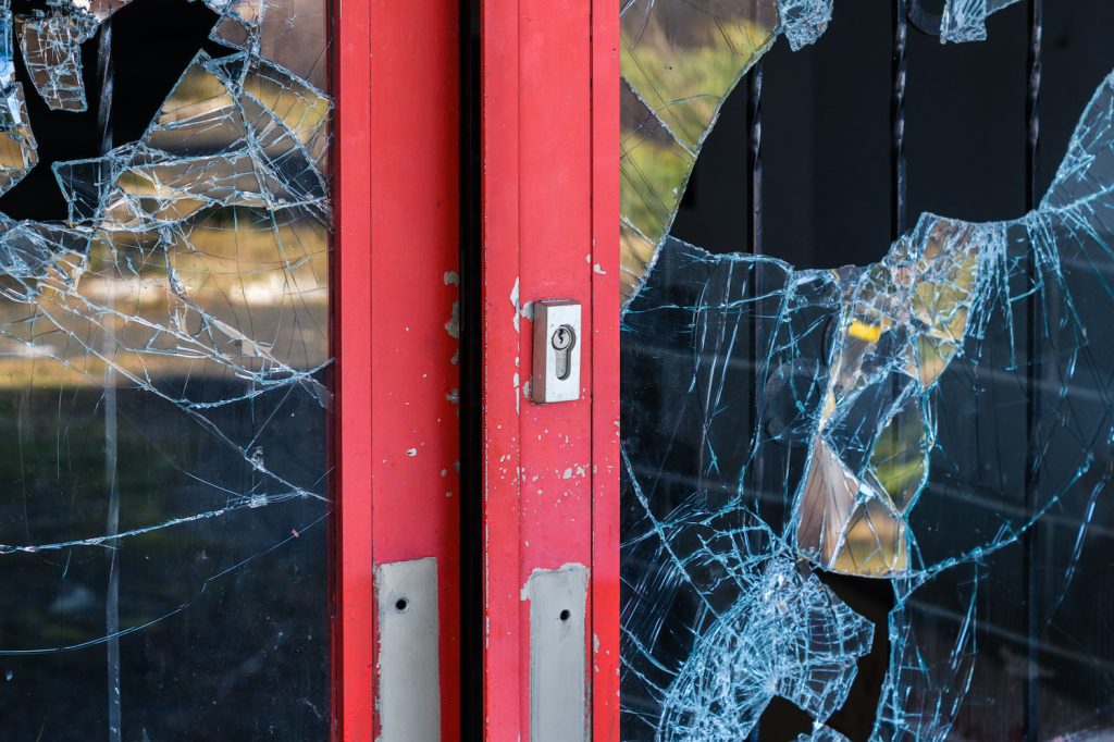 Close-up of a red door with shattered glass panels, showing signs of forced entry or vandalism.